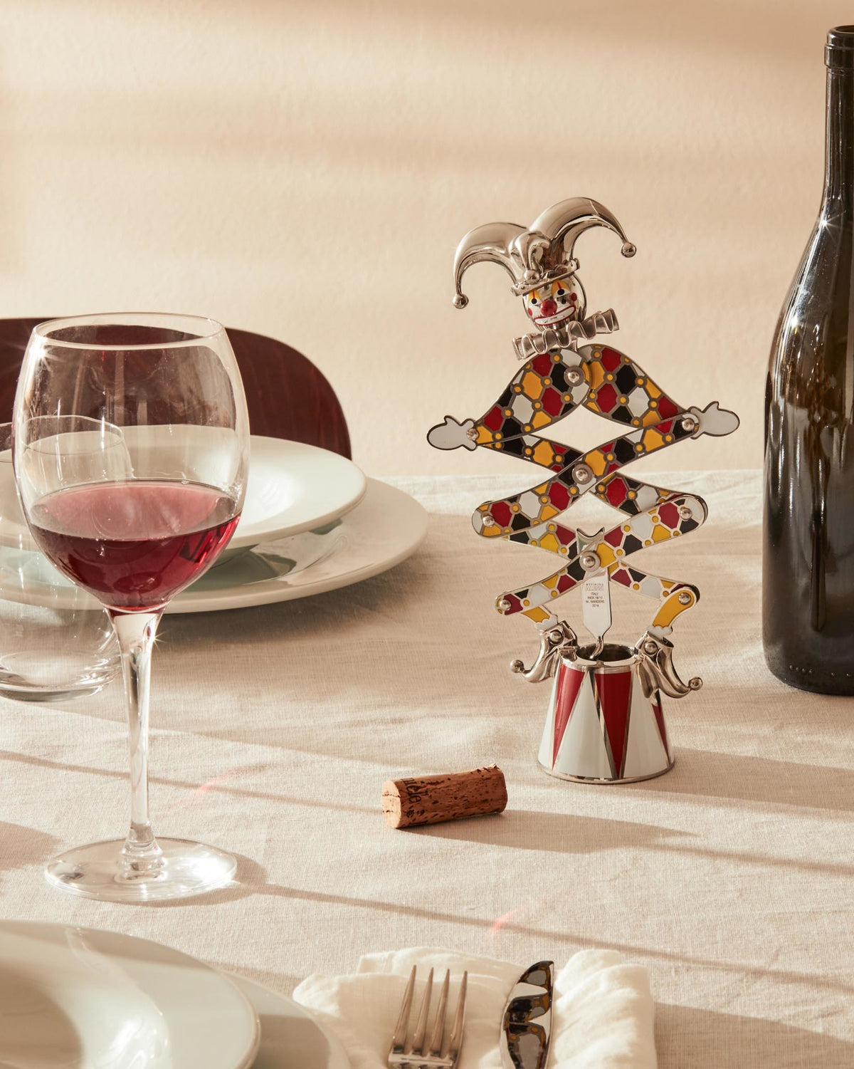 Jester-shaped corkscrew by Alessi, silver and multi-colored with a harlequin pattern, on a table next to a wine glass.