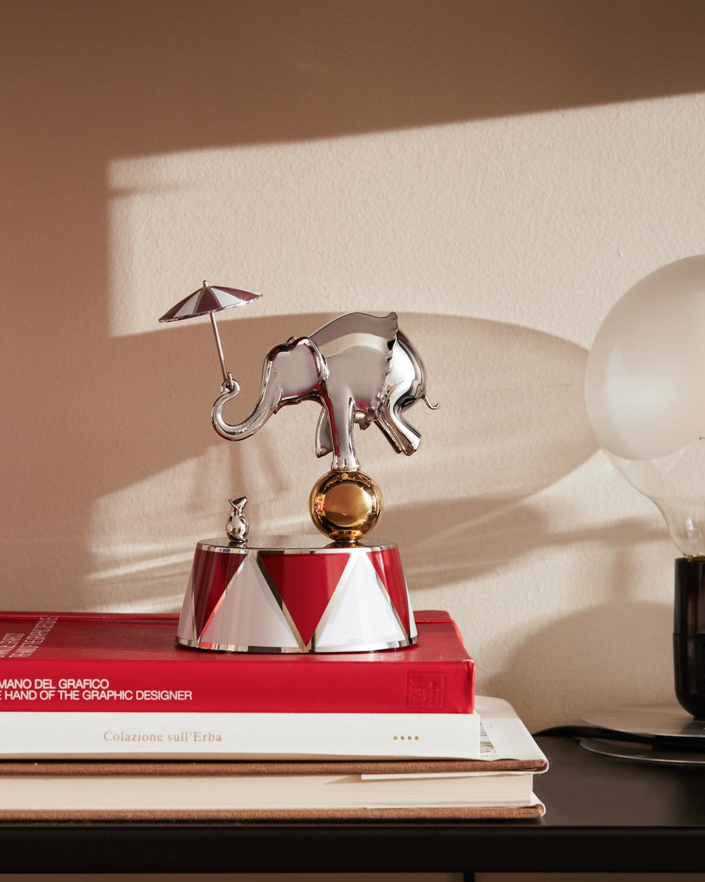 Chrome-plated elephant figurine by Alessi, balancing on a golden ball atop a red and white striped base, with a tiny mouse and umbrella.
