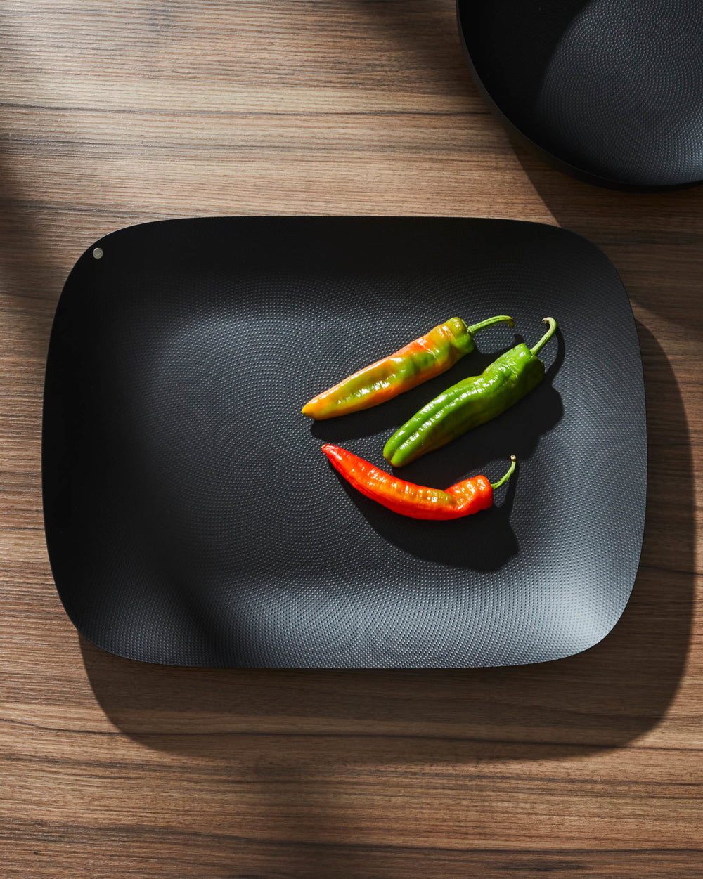 Black oblong serving tray by Alessi with a subtle dotted texture, holding three peppers on a wooden table.