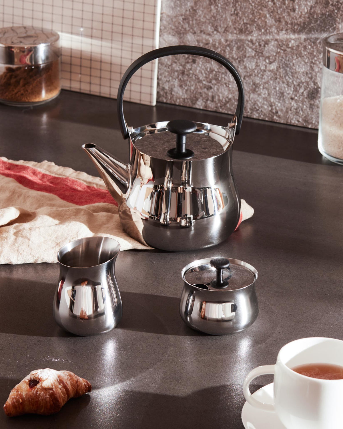 Polished stainless steel teapot by Alessi with black handle and lid knob, surrounded by matching serving pieces and breakfast items.