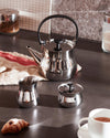 Stainless steel tea kettle by Alessi with a black handle, accompanied by matching milk and sugar vessels on a dark countertop.