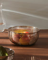 Polished metal slatted fruit bowl by Alessi, filled with colorful fruit on a wooden table.