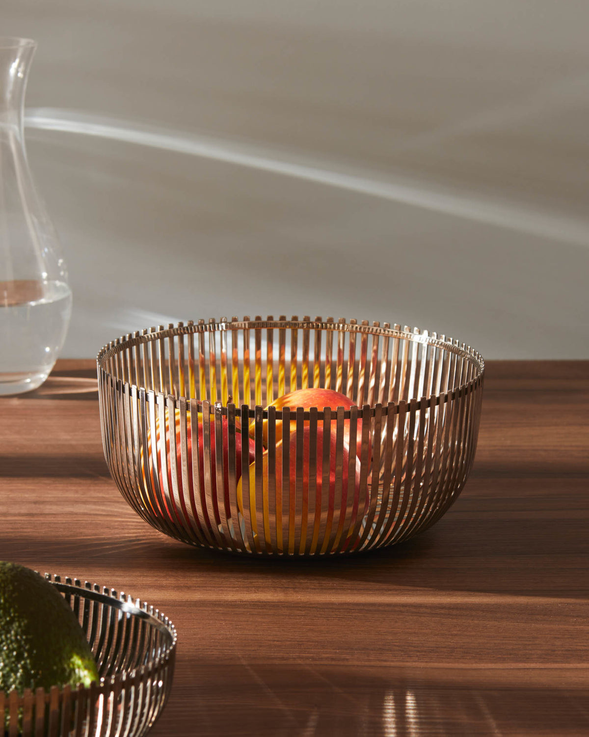 Polished metal slatted fruit bowl by Alessi, filled with colorful fruit on a wooden table.