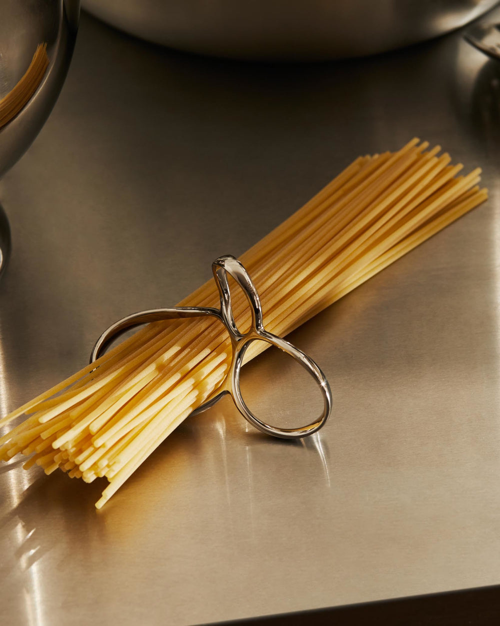 Silver-toned metal spaghetti measure by Alessi, holding a bundle of pasta on a brushed metal surface.