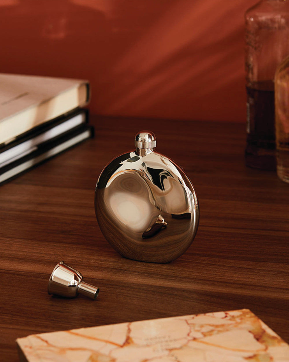 Shiny metal oval flask by Alessi, with screw cap and funnel, on a wooden table.