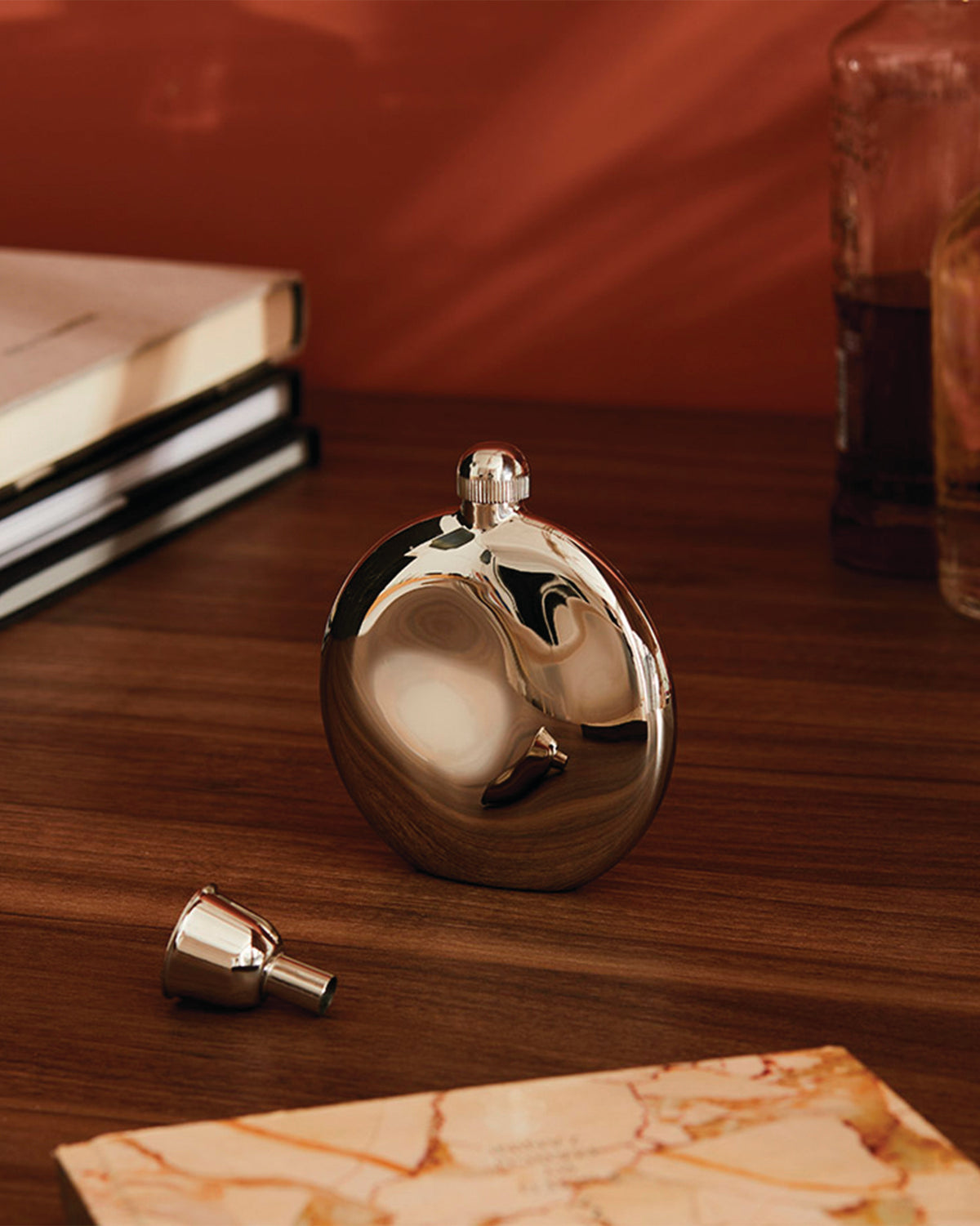 Shiny metal oval flask by Alessi, with screw cap and funnel, on a wooden table.