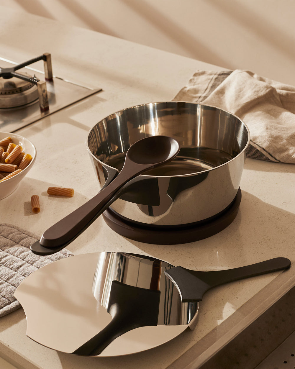 Polished stainless steel pasta pot by Alessi with a dark brown serving spoon and matching cooking tool, on a kitchen counter.