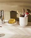 Birillo white bathroom accessories by Alessi, featuring a cylindrical toothbrush holder with toothbrushes and a soap dish with a yellow bar of soap on a sink.