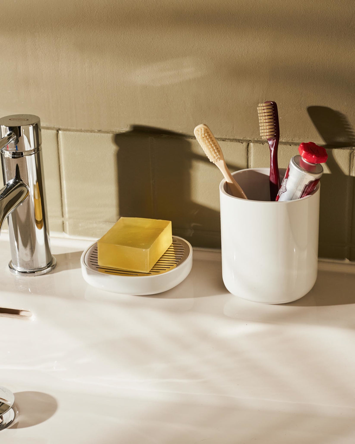 Birillo white bathroom accessories by Alessi, featuring a cylindrical toothbrush holder with toothbrushes and a soap dish with a yellow bar of soap on a sink.