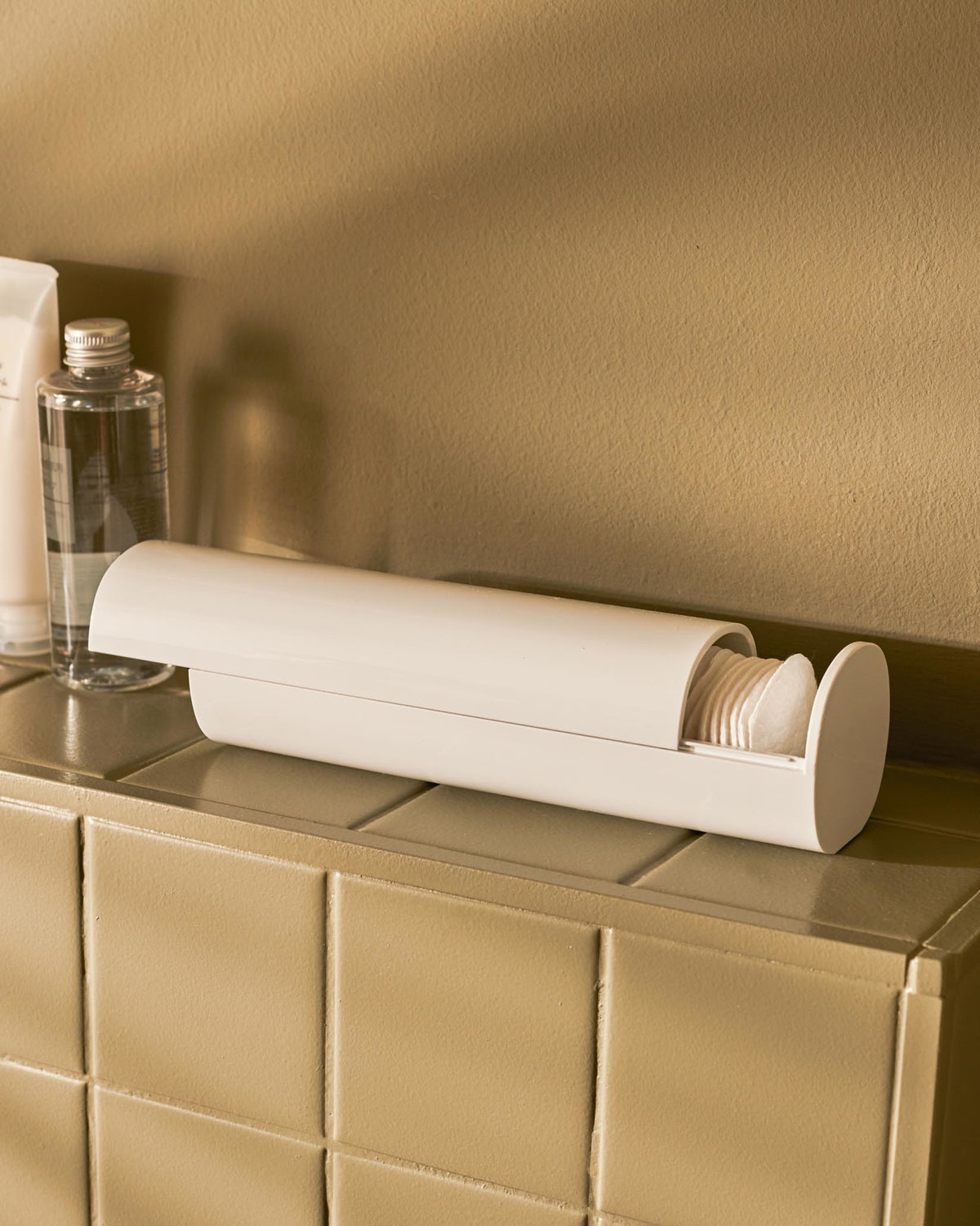 White cylindrical cotton pad dispenser by Alessi, open and filled with cotton pads on a beige tiled counter.
