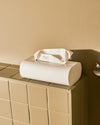 White tissue box holder by Alessi with curved ends and a white tissue emerging, on a beige tiled surface.