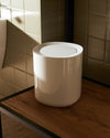 White cylindrical Birillo bin by Alessi with a round lid, placed on a wooden table in a tiled room.