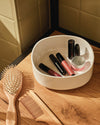 White oval Birillo container by Alessi holding lip glosses and a jar, beside a wooden brush and comb on a counter.