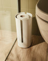 Off-white toilet paper holder by Alessi, holding white rolls, with a vertical slot, on a light wood floor.