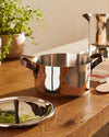 Stainless steel casserole pot by Alessi with brown leather handles on a rustic wooden table, lid nearby.