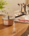 Stainless steel saucepan by Alessi with a dark brown handle and lid knob, on a wooden counter with a striped tea towel.