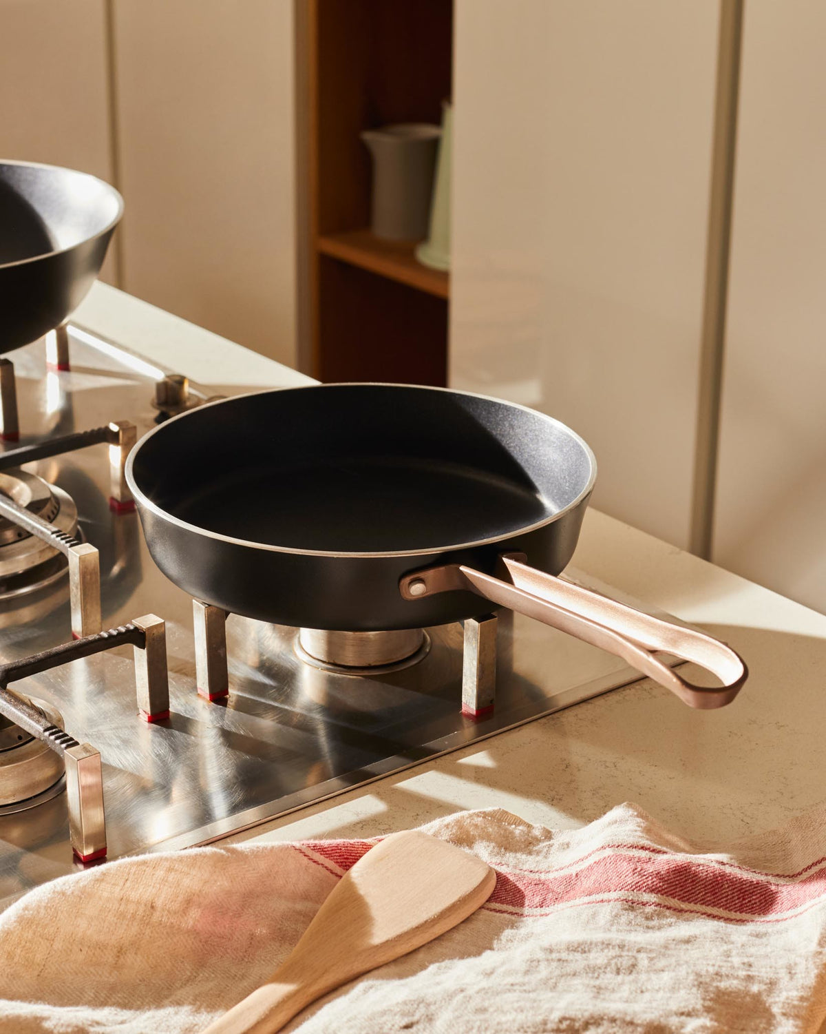 Black frying pan with rose gold handle by Alessi, resting on a stainless steel stovetop in a sunlit kitchen.
