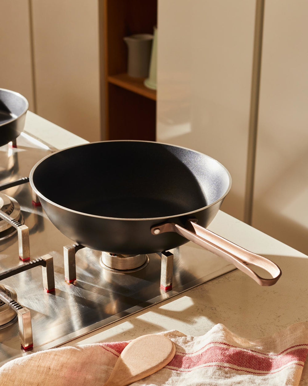 A black non-stick pan by Alessi with a rose gold handle, sitting on a gas stove next to a striped kitchen towel and wooden spoon.