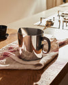 Shiny metallic pitcher with a dark handle by Alessi, resting on a striped towel on a wooden counter.