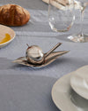 Silver metal apple-shaped oil cruet by Alessi, on a sleek leaf tray with a long dipper, on a light blue tablecloth.