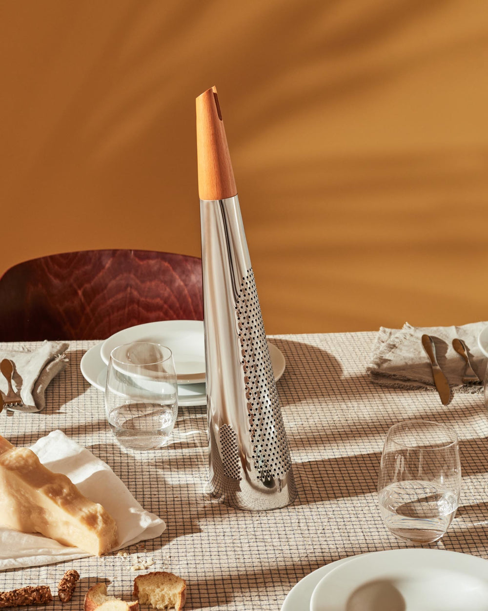 Conical stainless steel grater by Alessi with a light wood handle on a checkered tablecloth.