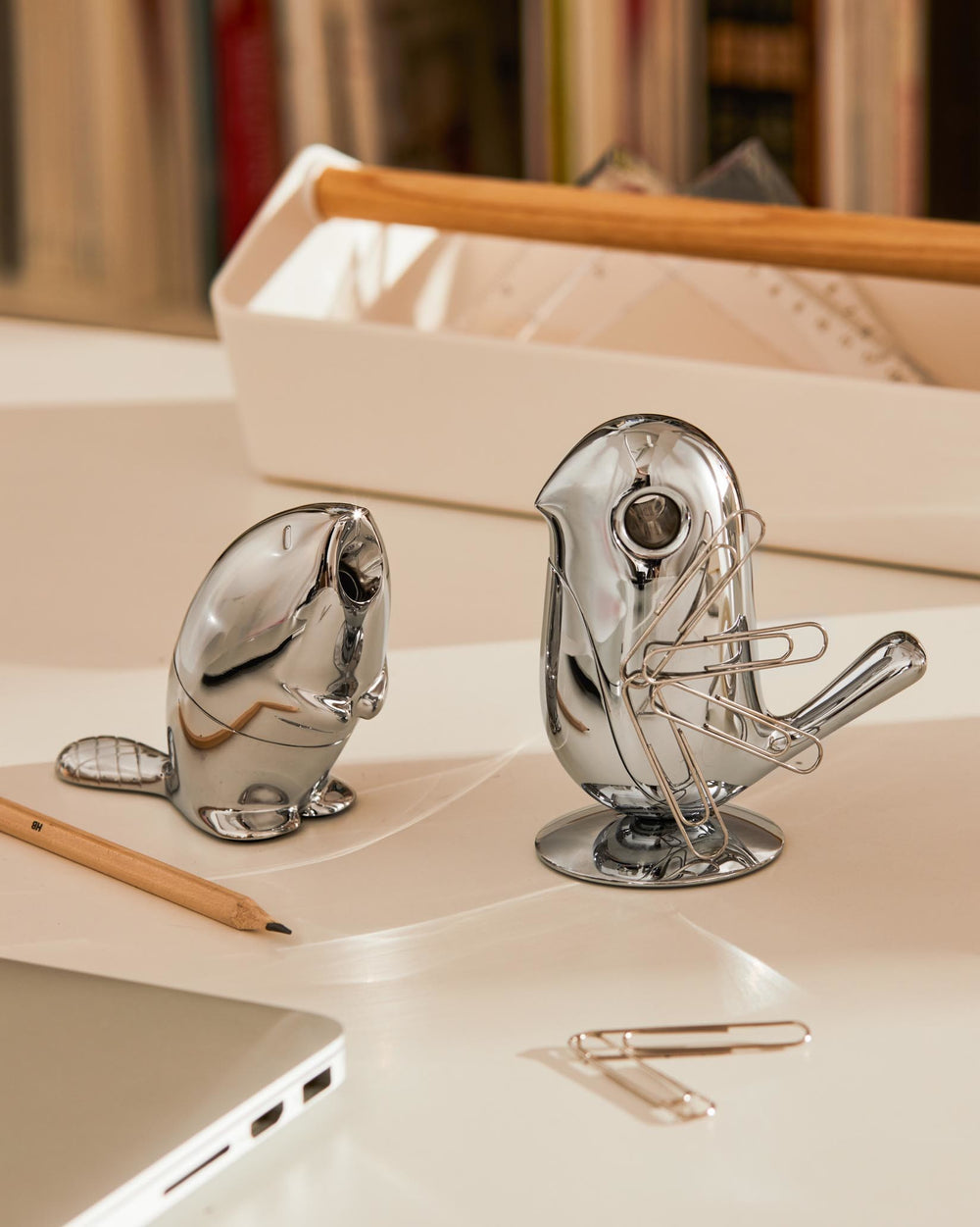 Chrome-plated beaver pencil sharpener by Alessi, with a wooden pencil and bird paperclip holder on a desk.