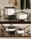Mami polished stainless steel pot set by Alessi, gleaming on two metal kitchen shelves with utensils.