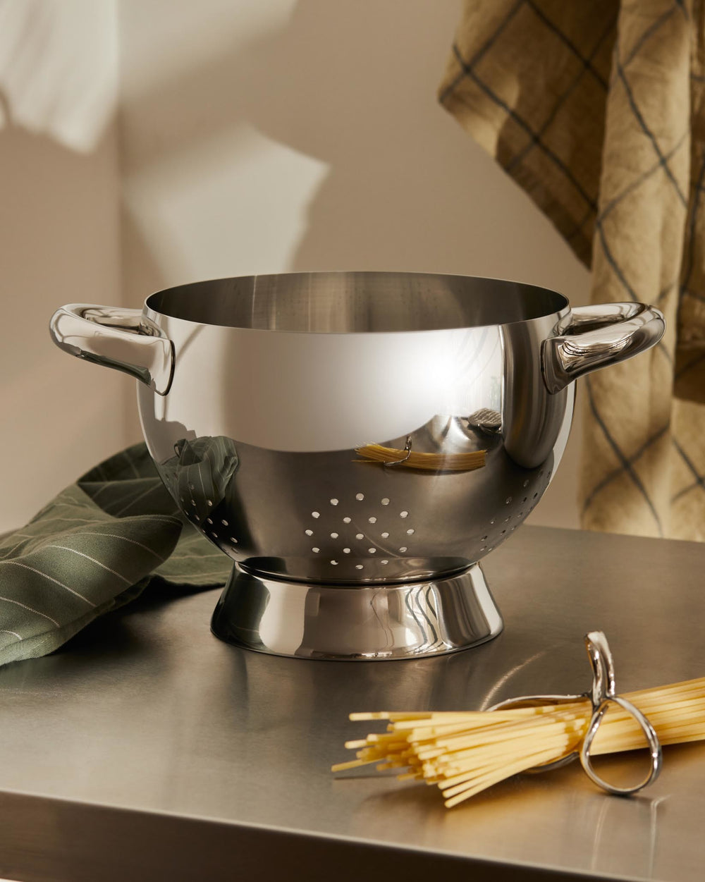 Polished stainless steel colander by Alessi with handles, perforated base, on a metal countertop with spaghetti and a green towel.