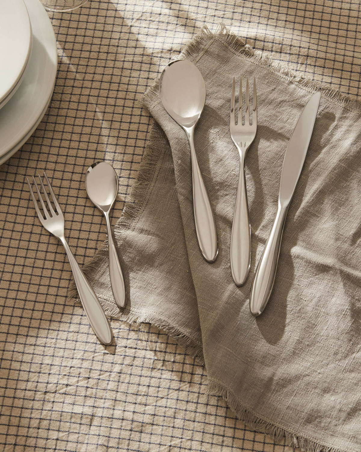 Mami silver cutlery set by Alessi on a beige plaid tablecloth with a fringed napkin.