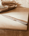 Silver metallic knife by Alessi, with an ergonomic handle, angled on a light marble cutting board and a brown checkered cloth with frayed edges.