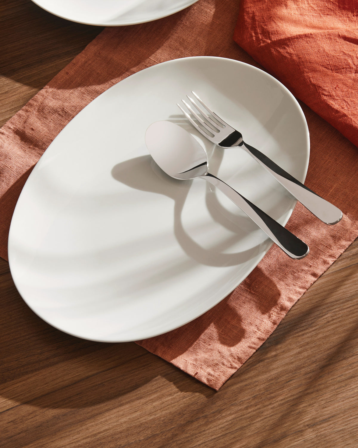 White oval plate with sleek silver cutlery by Alessi, resting on a rust-colored placemat on a wooden table.