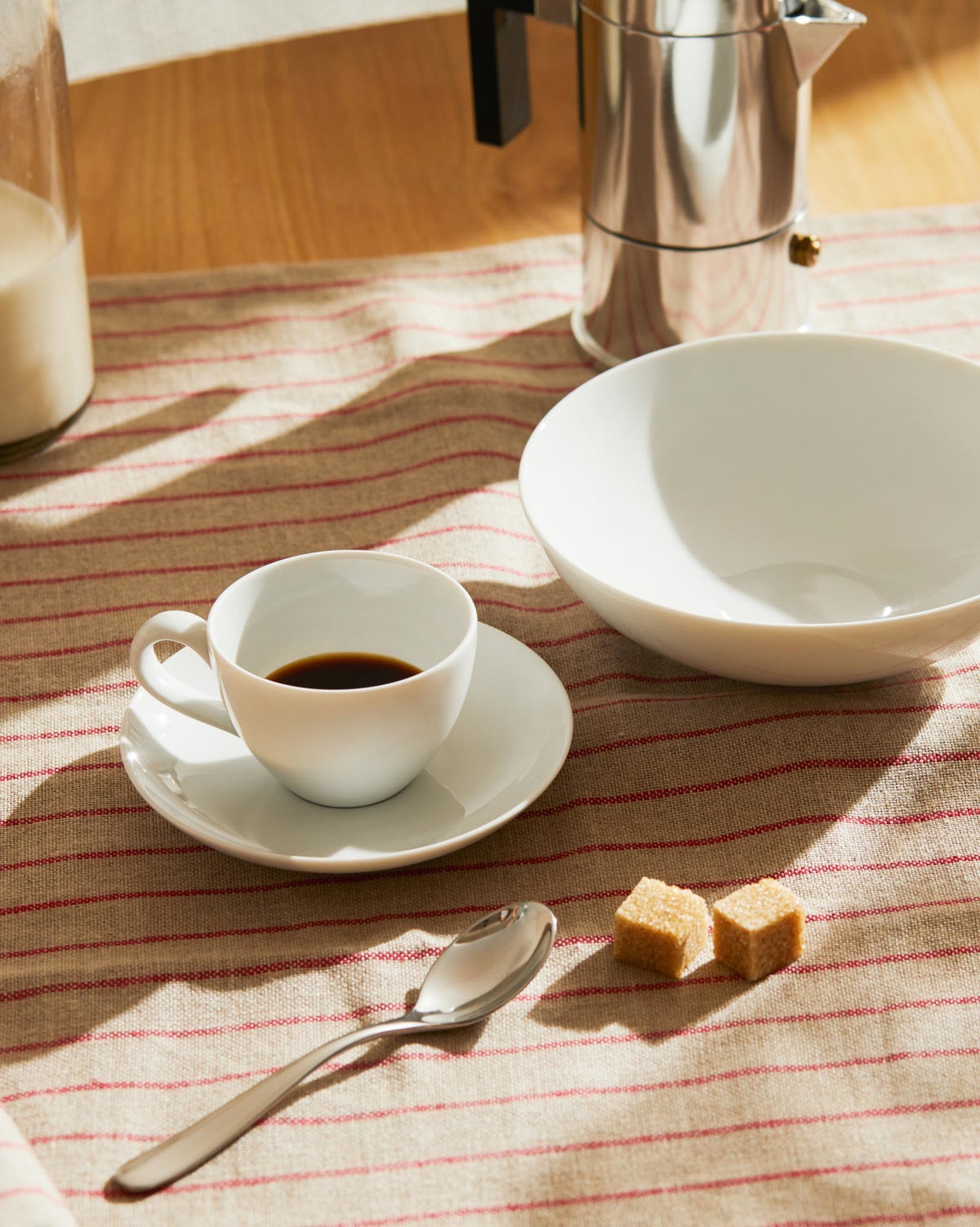 Stainless steel Moka pot by Alessi with black handle, shown on a striped tablecloth with a white coffee cup and bowl.