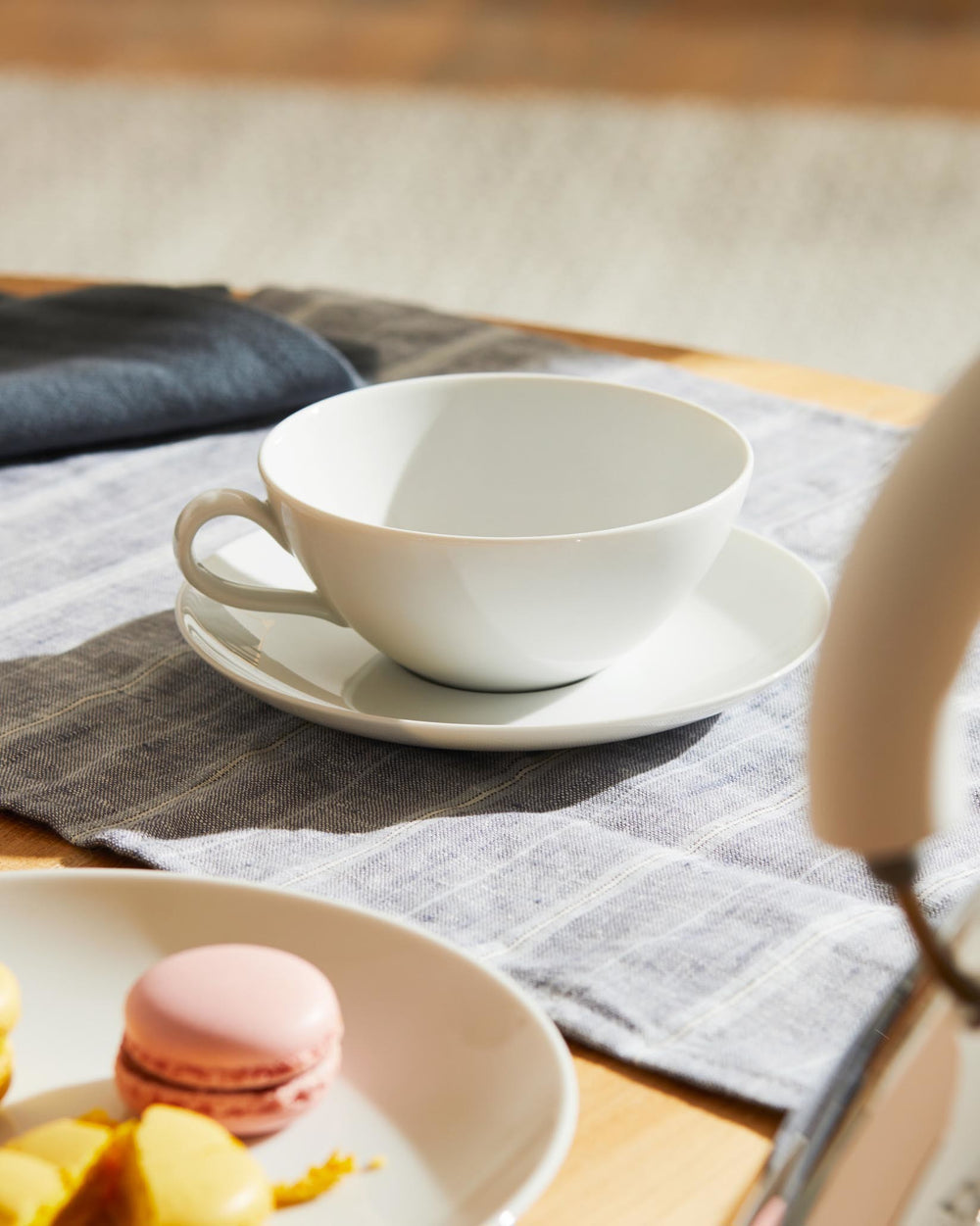 White ceramic cup and saucer by Alessi on a grey striped placemat, next to a plate with a pink macaron and yellow dessert pieces.