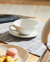 White ceramic teacup and saucer by Alessi on a striped grey placemat with macarons and pastries.