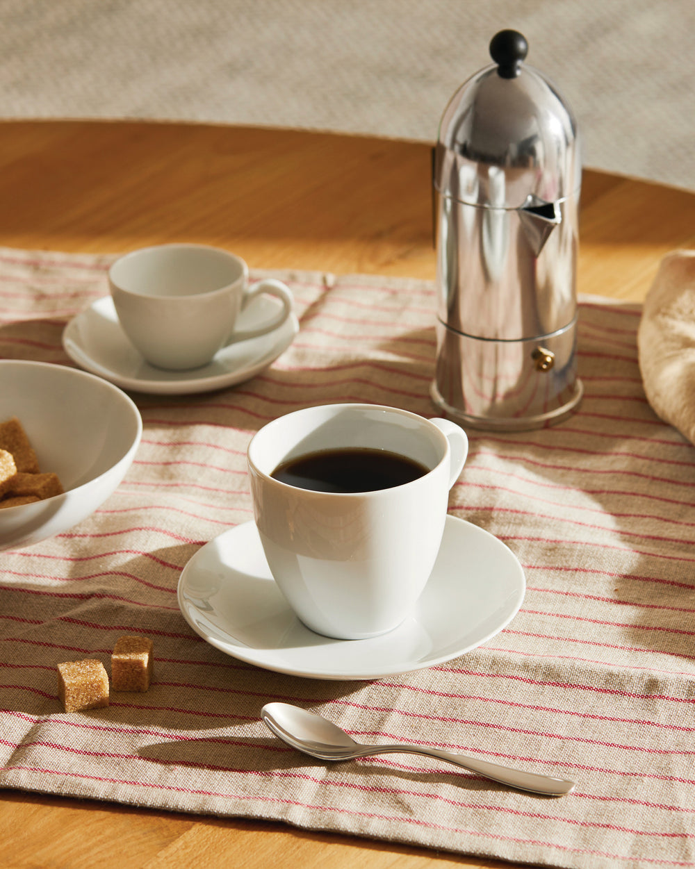 Silver Mami espresso maker by Alessi, styled with white cups, brown sugar, and a spoon on a striped cloth.