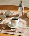 Mami metallic coffee maker by Alessi, on a red-striped linen placemat with white coffee cups and sugar cubes.