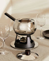 Chrome fondue set by Alessi with a black handle, textured black stand, and separate lid with a dark wood knob, on a light linen tablecloth.