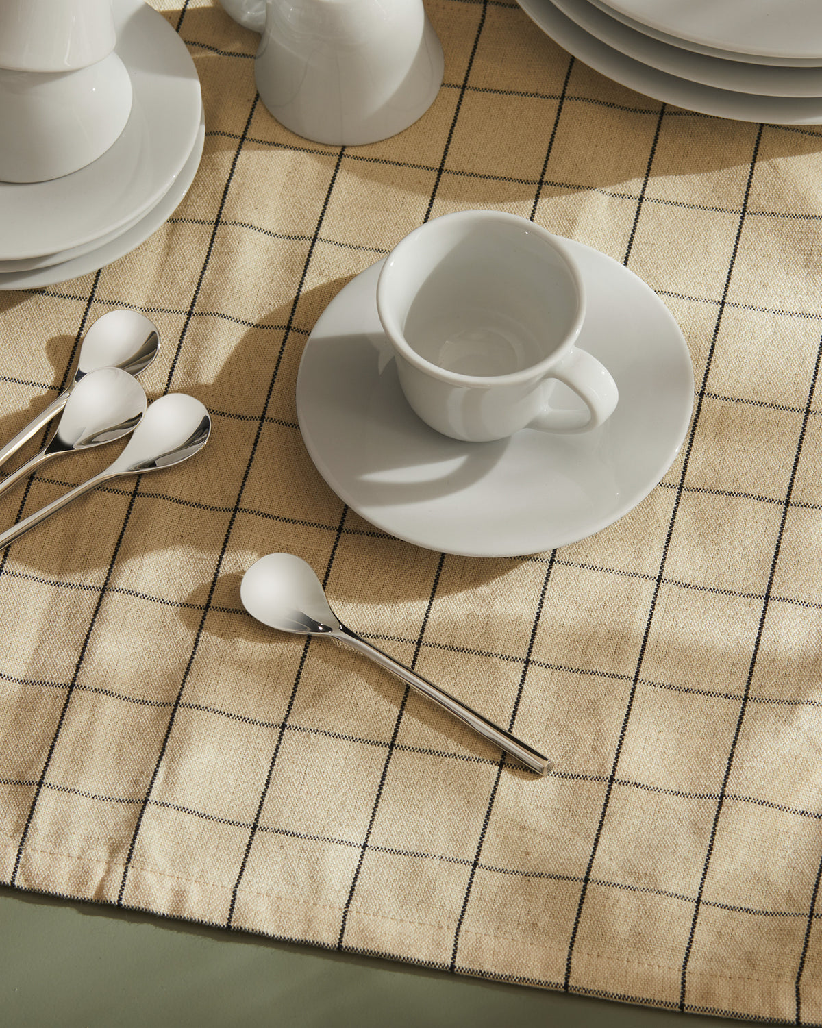 White ceramic dishes by Alessi displayed with cups and silver spoons on a beige plaid tablecloth.