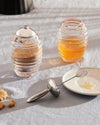 Glass ribbed honey pot by Alessi, filled with honey, with a dipper and matching sugar pot on linen.