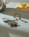 Silver metal tea infuser by Alessi with a chain, alongside loose tea leaves, a silver teapot, and lemon slices.