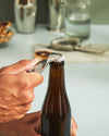 Silver metallic bottle opener by Alessi opening a brown beer bottle held by a hand.