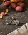 Silver potato peeler by Alessi, with a modern curved design, on a dark countertop with red potatoes.