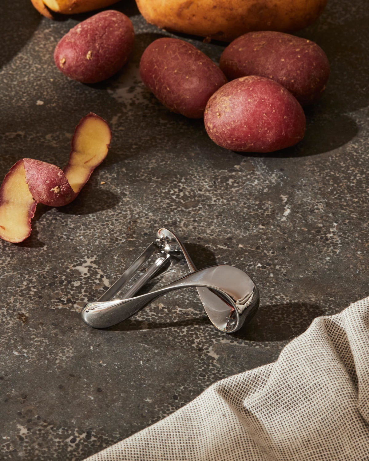 Silver potato peeler by Alessi, with a modern curved design, on a dark countertop with red potatoes.