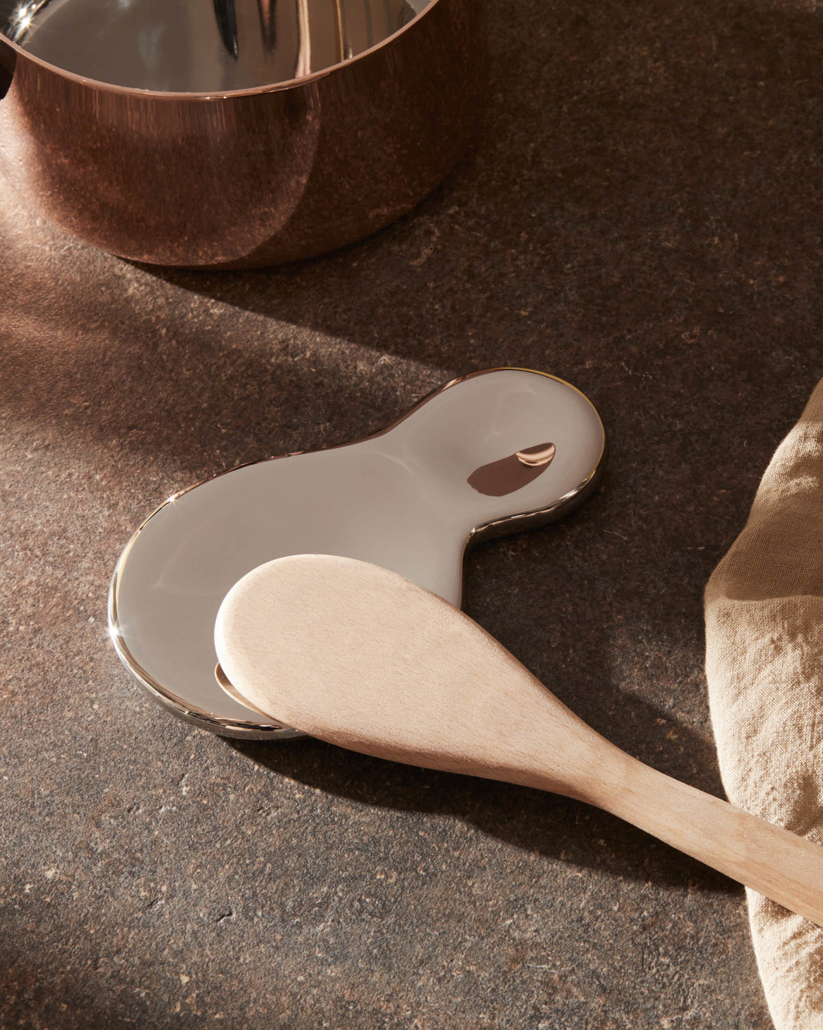 Organic-shaped, glossy silver-toned spoon rest by Alessi, with a wooden spoon on a dark stone surface.