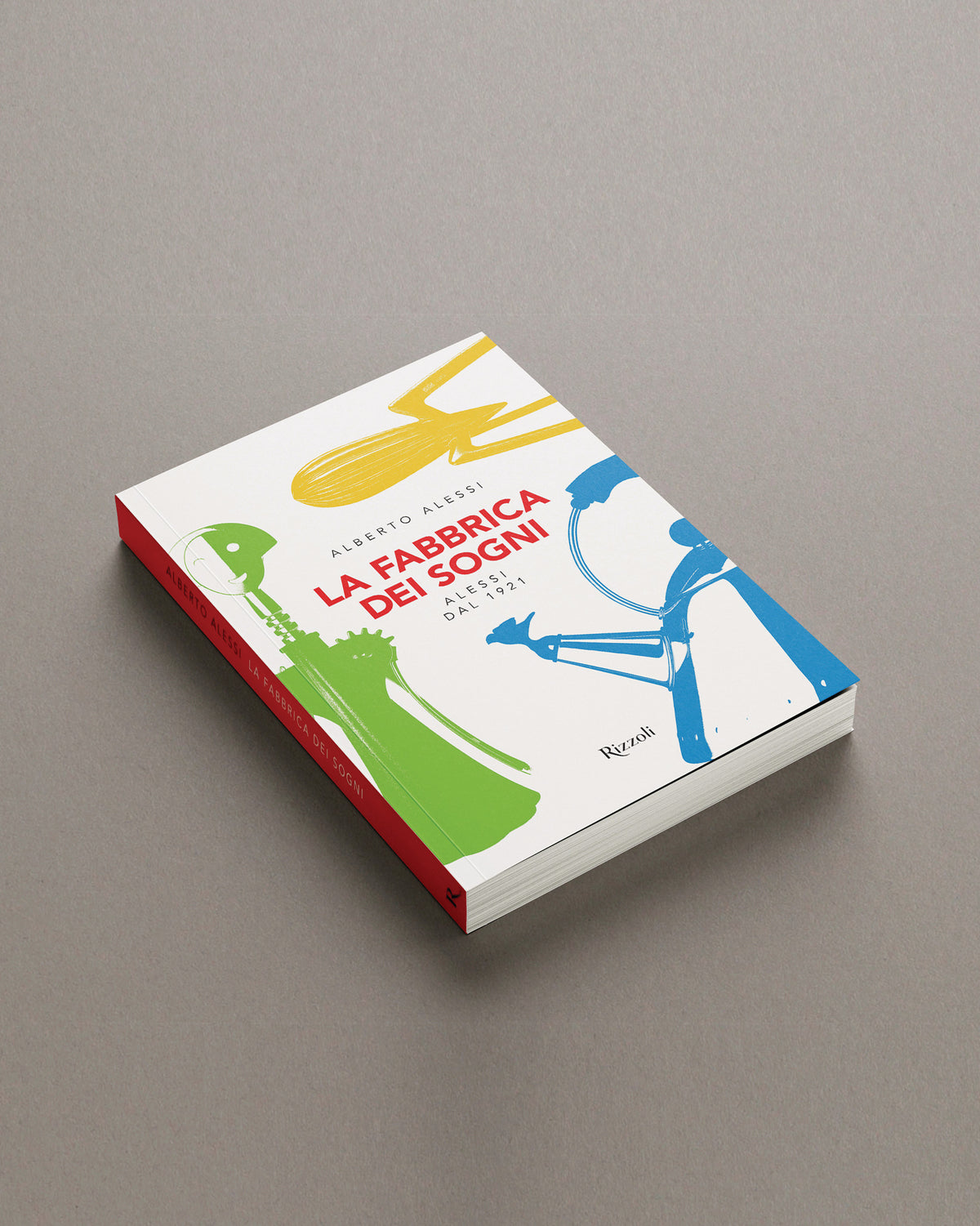 A white hardcover book titled 'La Fabbrica dei Sogni' by Alessi, with yellow, green, and blue kitchenware illustrations, displayed at an angle on a grey surface.