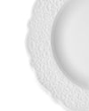 White ceramic plate by Alessi with a scalloped edge and embossed scrollwork pattern, partially visible.