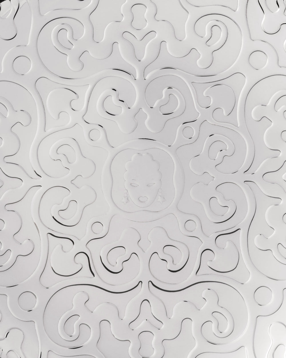 White embossed pattern by Alessi featuring swirling, floral-like designs and a central medallion with a stylized face wearing star earrings.