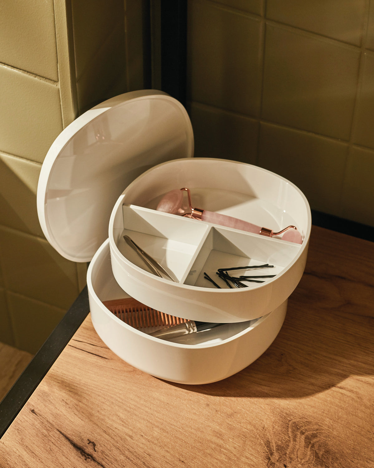 White oval stacked organizer by Alessi, with a rose quartz facial roller, tweezers, bobby pins, and a wooden comb.