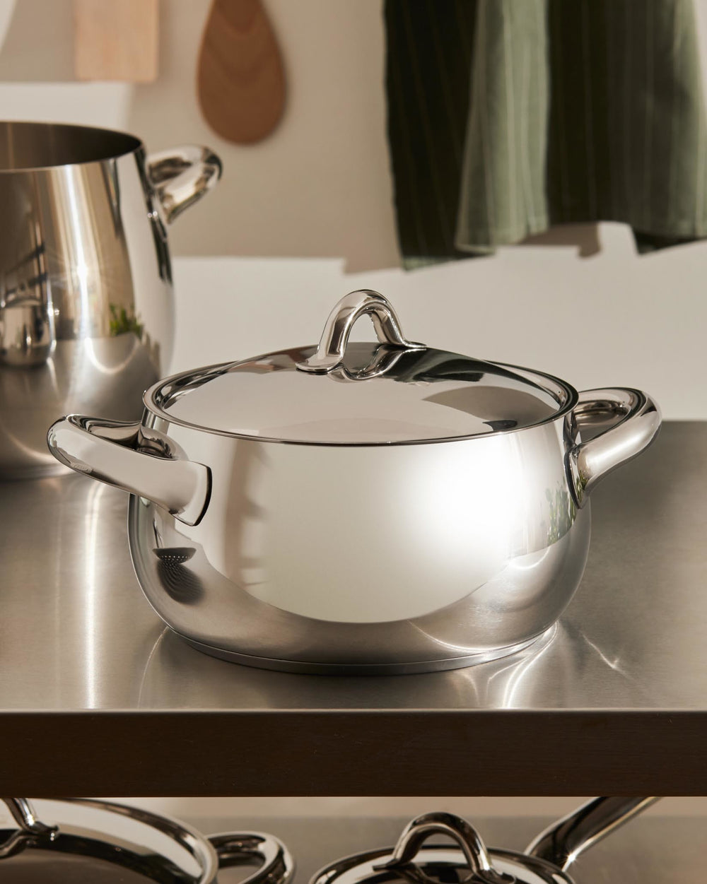 Polished stainless steel cooking pot by Alessi, with a lid and two handles on a metallic counter.