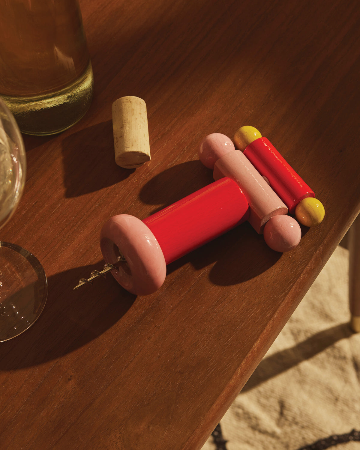 Red and pink ES17 corkscrew by Alessi, featuring yellow accents, on a wooden table with a wine bottle and cork.
